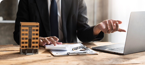 Persona trabajando en una mesa con una maqueta de edificio, documentos y una computadora portátil.