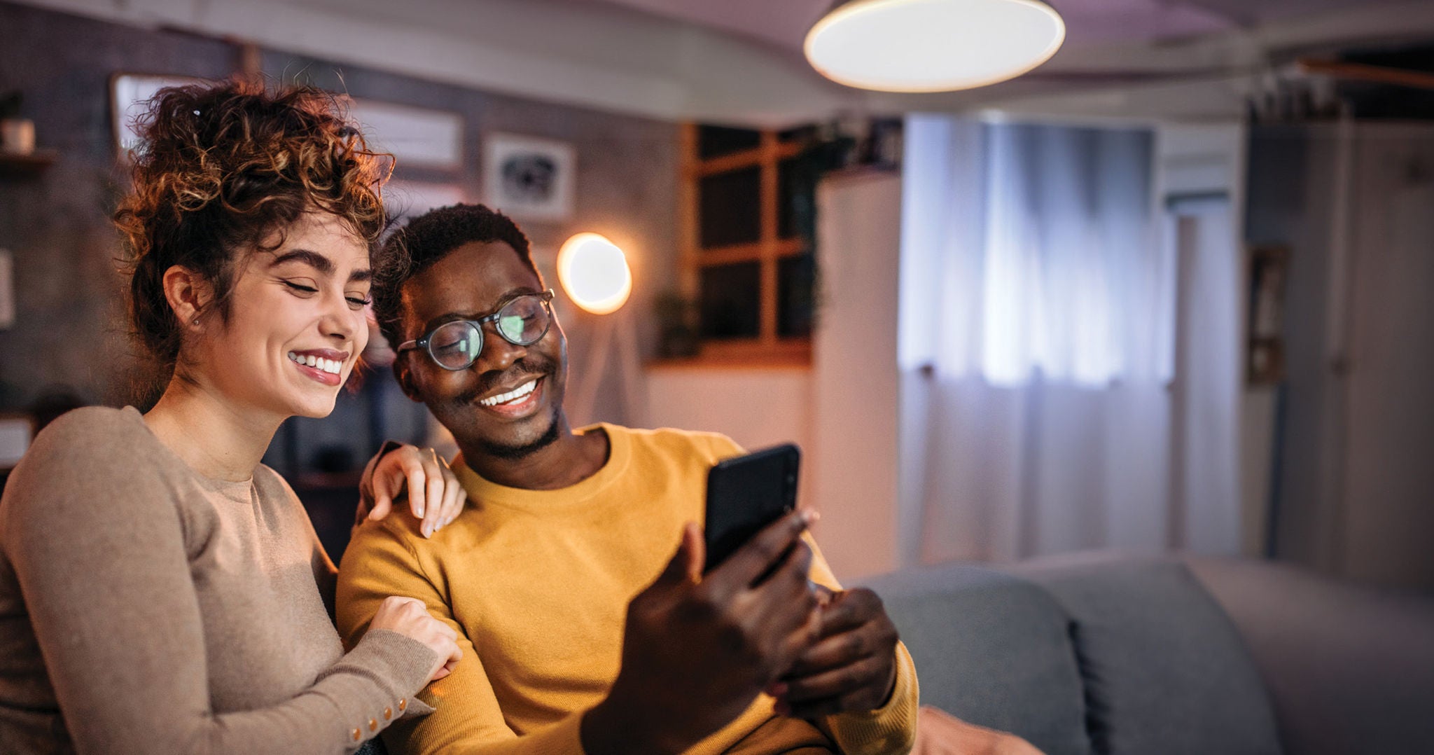Two people sitting close together on a couch, looking at a smartphone in a warmly lit living room.