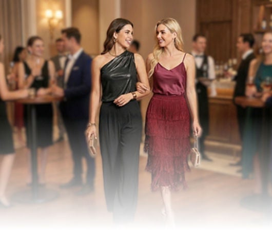 Dos personas en un evento elegante, usando conjuntos de moda: un jumpsuit negro de un solo hombro y una falda de volantes con blusa satinada.