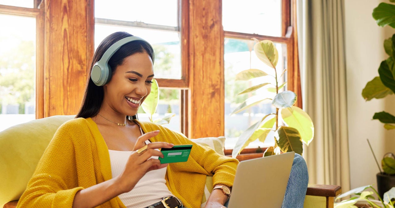 Mujer sentada en sofa con tarjeta de debito en mano y utilizando una computadora portátil