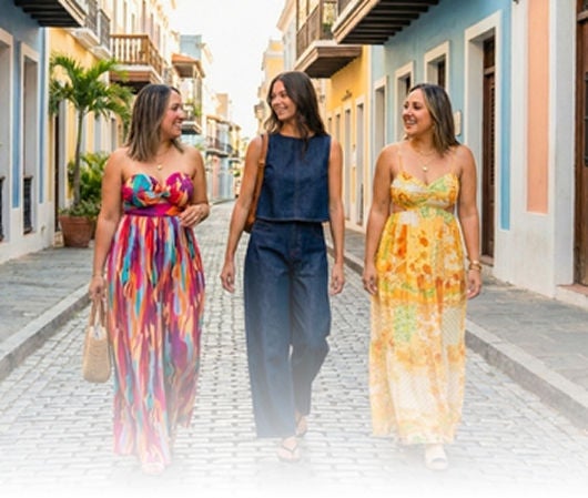 Tres mujeres caminando por calle adoquinada con vestidos coloridos y azul. 